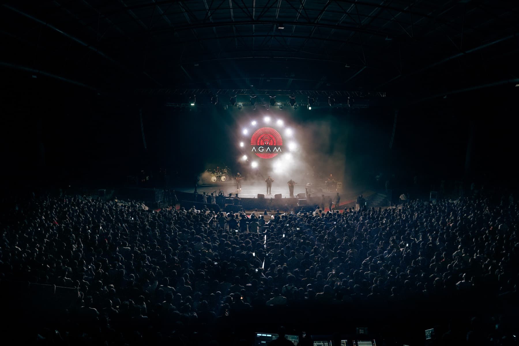 Wide arena view of Agam performing before a packed audience with the band logo lit on stage